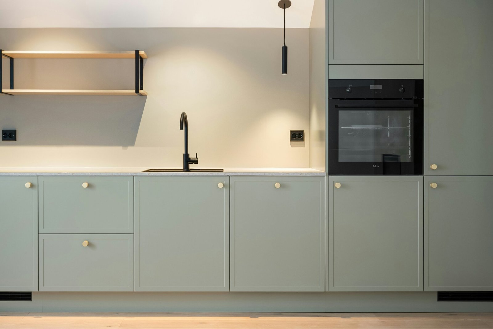 Modern kitchen with soft sage green cabinets, minimalist black fixtures, and warm neutral lighting showcasing 2026 earthy green cabinet color trends, photo by Lisa Anna.