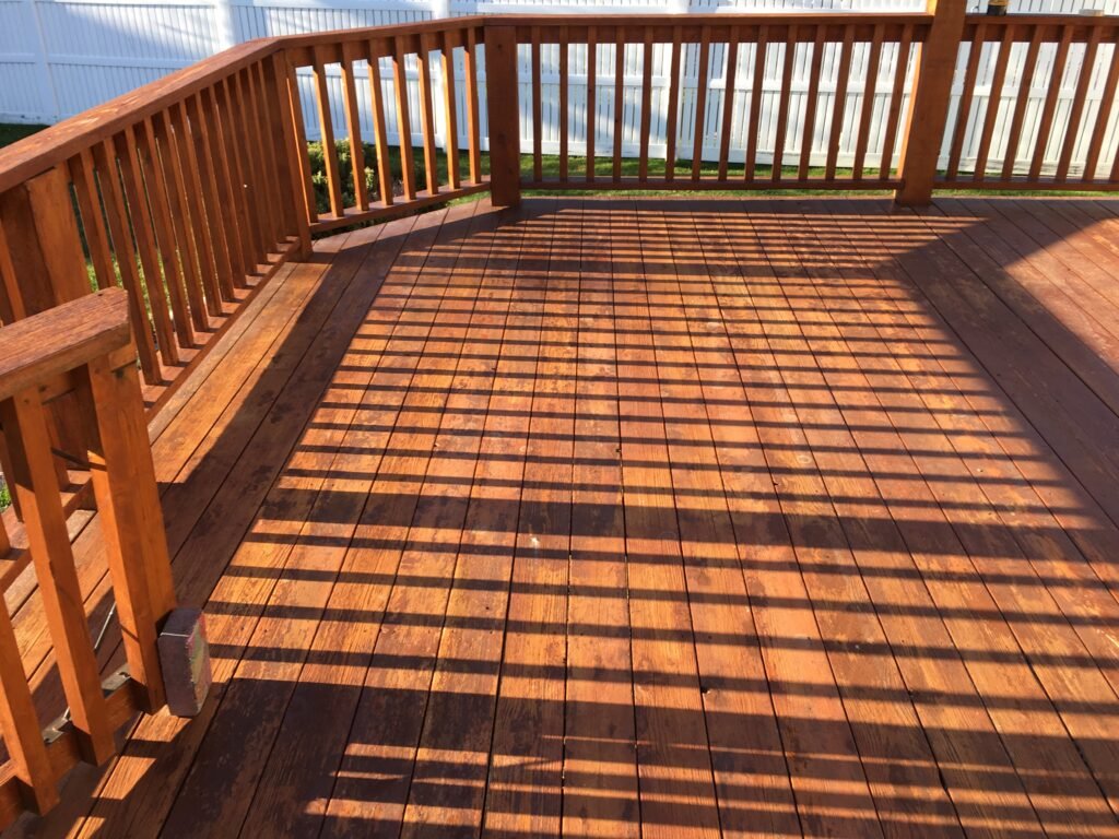 Freshly stained wooden backyard deck completed by Paint EZ, featuring a rich brown finish, clean railings, and sunlight casting striped shadows across the deck boards.