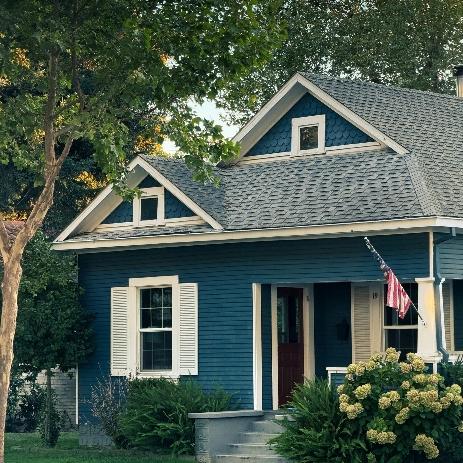 Traditional blue house with white shutters, a gray roof, and an American flag